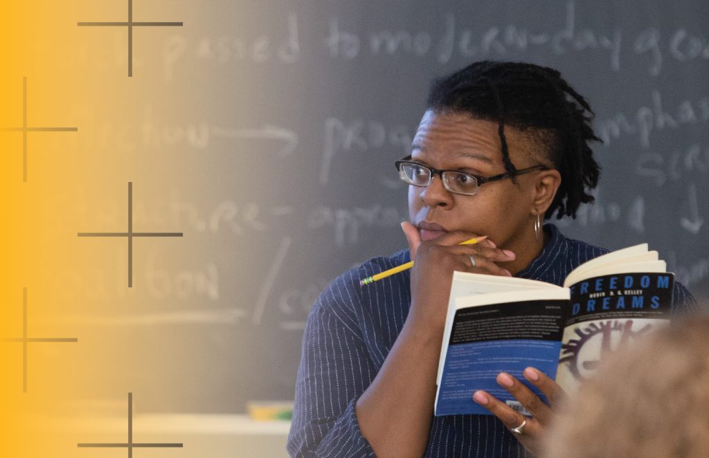 Professor with a book and a chalkboard behind her.