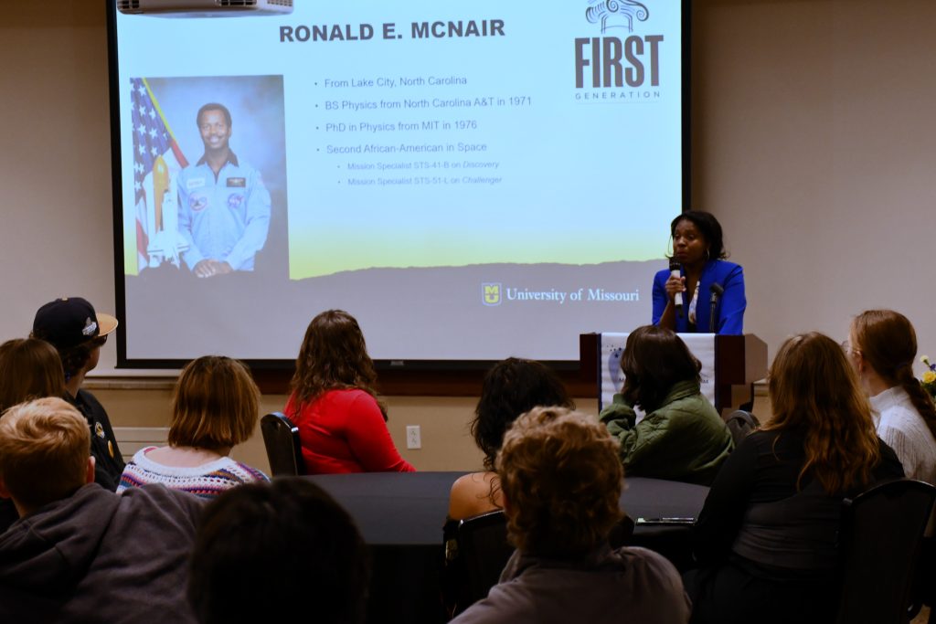 Dr. Natalie Downer stands in front of a projector image of Ronald McNair with a text bio, speaking to a room of people