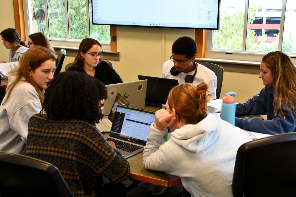 six students on their laptops around a table collaborating on the research project