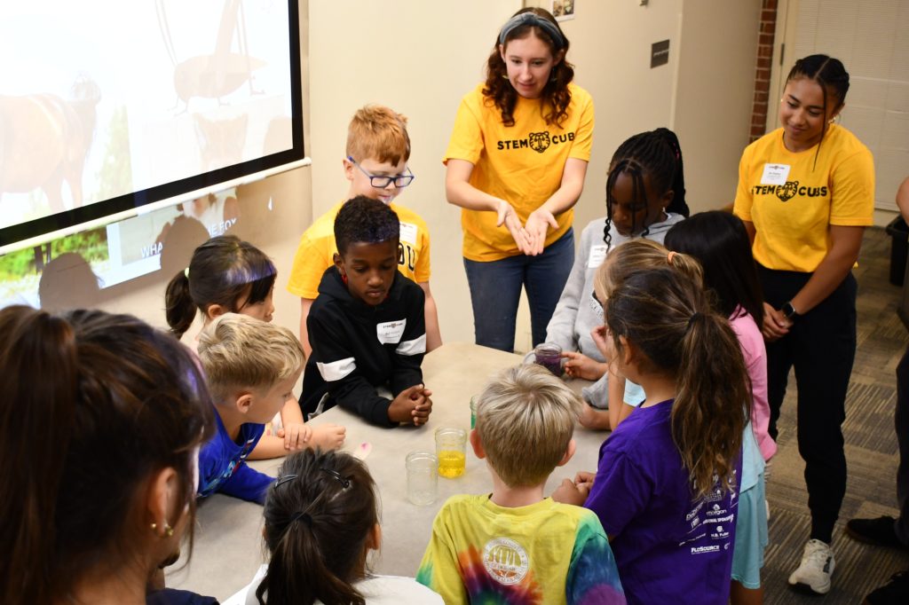 Grades K-2 classroom gathers around a table of liquid-filled glasses during a lesson on waves and sound