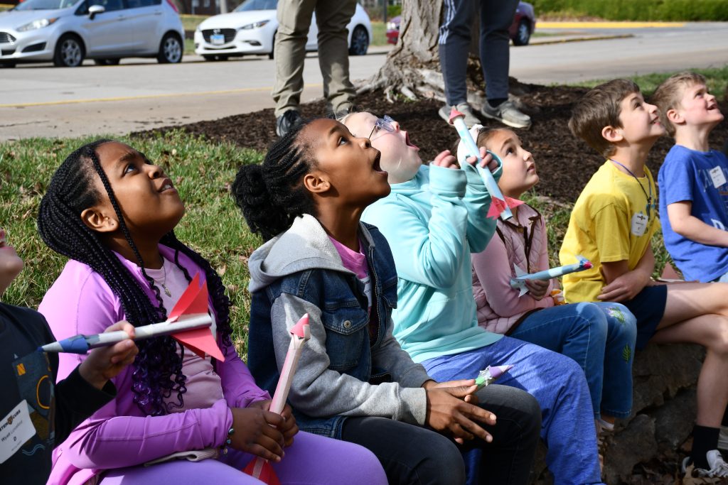 a row of elementary aged children sit in the grass and look up as their rockets launch