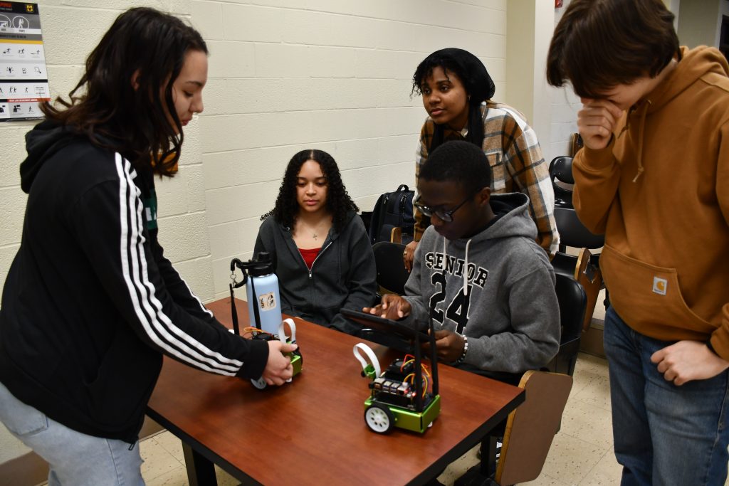 a small group of college students use a tablet device to control two small vehicle robots