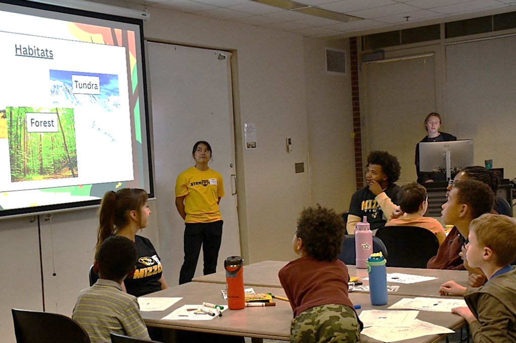 students at tables face projector screen and female presenter in classroom